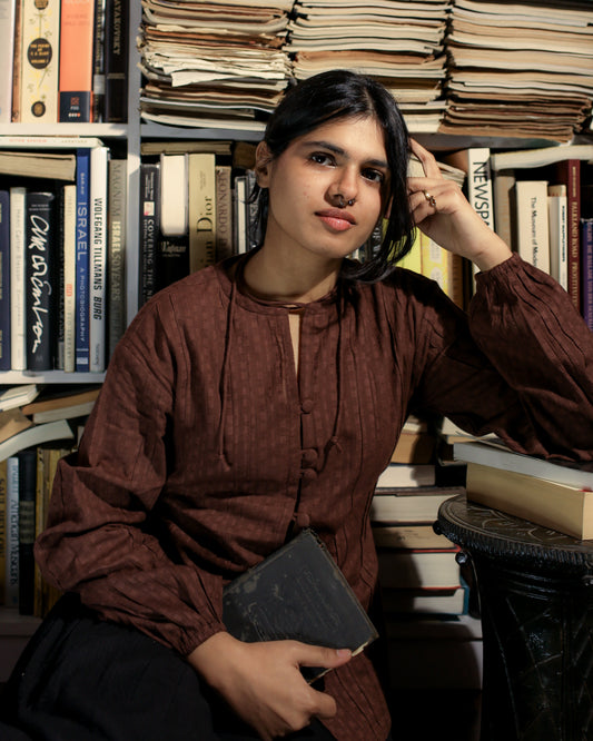 Woman wearing chocolate brown cotton dobby shirt with pintucks and balloon sleeves, styled with a book in a cozy reading nook.