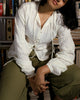 Model wearing ivory white cotton dobby shirt with pintucks and balloon sleeves, seated in front of bookshelf.