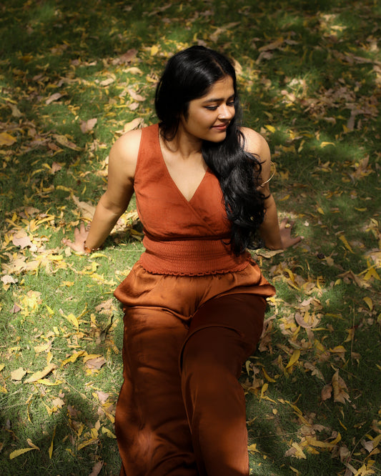 Woman in burnt ochre sleeveless V-neck cotton top with smocked waist, sitting on grass surrounded by autumn leaves.