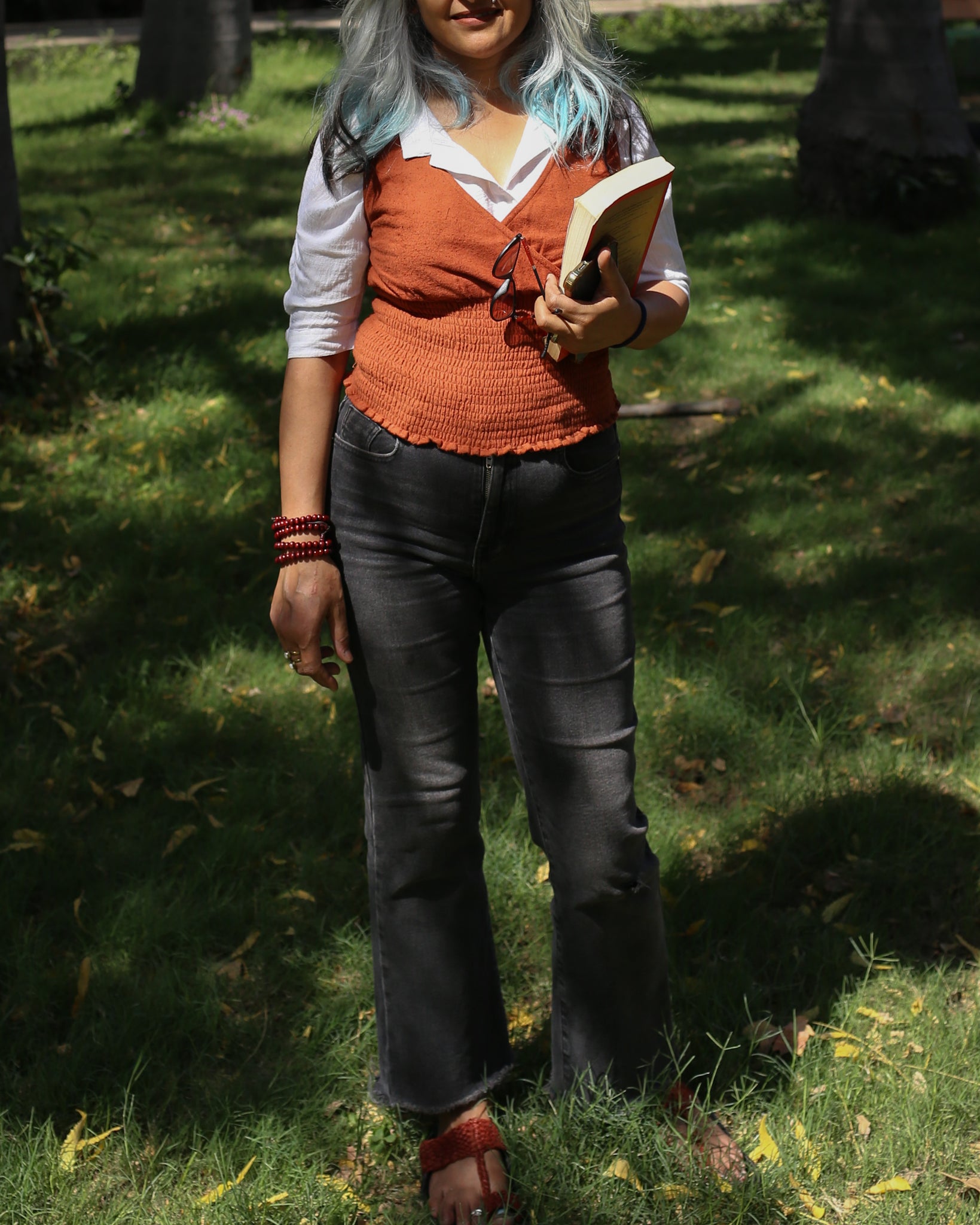 Burnt ochre sleeveless top layered with shirt and paired with grey pants, holding a book outdoors.