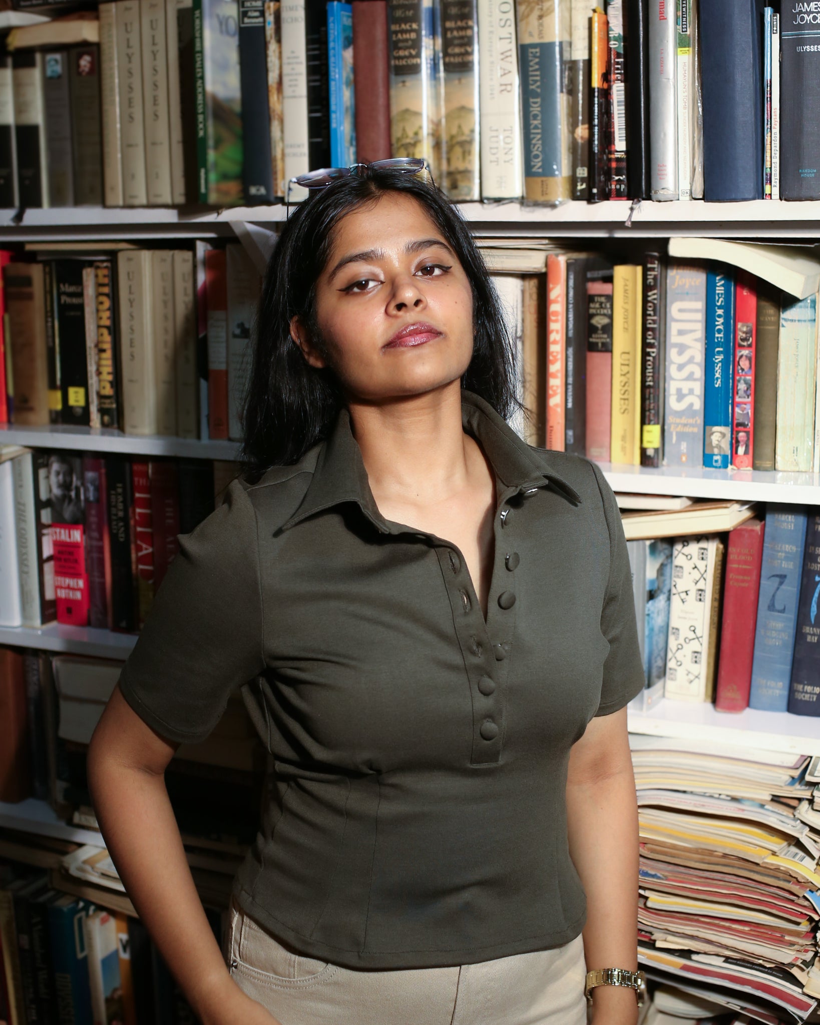 Woman wearing polo top having fullness in chest standing in front of a bookshelf filled with books